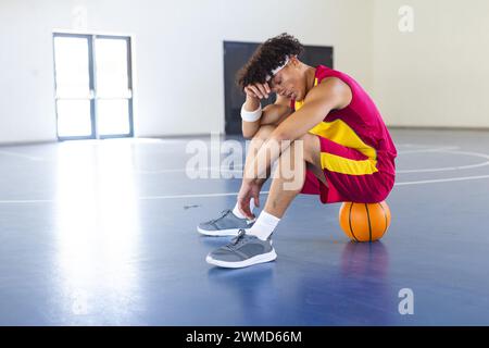 A young biracial man sits on a basketball, looking thoughtful Stock Photo