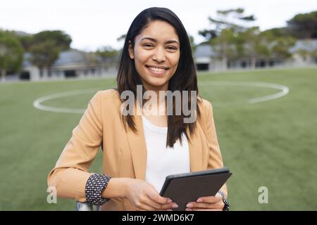 Young biracial teacher smiles holding a tablet on a sports field Stock Photo