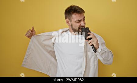 Joyful young hispanic man, confidently wielding a brush as a mic, belting his heart out in a song isolated on a yellow background Stock Photo