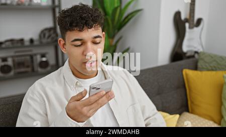 A handsome young man focuses intently on his smartphone while sitting comfortably in a modern living room. Stock Photo