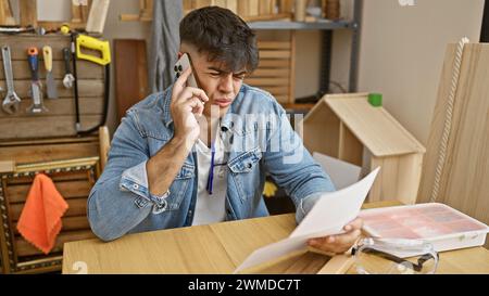 Handsome young hispanic man, a skilled carpenter, engrossed in reading a document while talking on his smartphone at his carpentry workshop Stock Photo