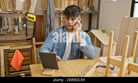 Hardworking young hispanic man, a dedicated builder and carpenter engaged in his profession, talking on smartphone and using touchpad amidst the lumbe Stock Photo