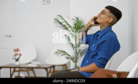 Handsome young latin man, tattooed worker engrossed in lively conversation on his cellphone, sitting in indoor chair, laughing with a business profess Stock Photo