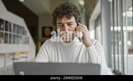 A focused hispanic man with a beard works indoors at his laptop while talking on his phone in an office setting. Stock Photo