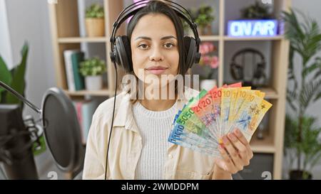 Hispanic woman with headphones in radio studio holding swiss francs, projecting confidence and professionalism. Stock Photo