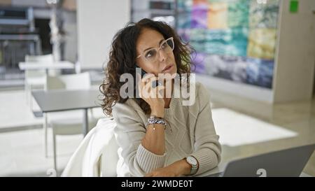 A thoughtful middle-aged hispanic woman converses on a smartphone in a modern office setting, evoking professionalism and engagement. Stock Photo