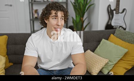 A smiling young hispanic man with curly hair sits casually at home on a grey couch, exuding a relaxed and handsome aura. Stock Photo
