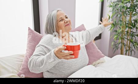 A smiling senior woman holding a red mug, stretching in bed with pink pillows and a plant in a cozy bedroom Stock Photo