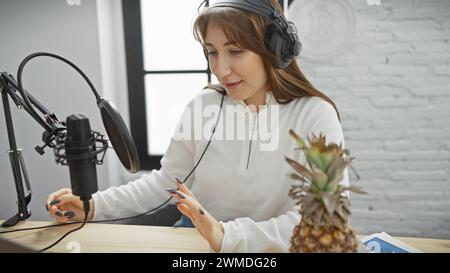 A young woman with headphones speaks into a microphone in a podcast studio setup with a casual tone. Stock Photo