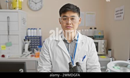A young asian man wearing safety goggles and a lab coat stands confidently in a clinical laboratory setting. Stock Photo