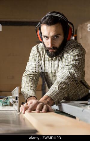 Vertical photo a portrait of a focused carpenter man mid adult caucasian, equipped with safety ear muffs, as he carefully pushes timber through a tabl Stock Photo