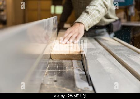Horizontal photo close-up of a woodworker's hands as they guide a piece of wood through a table saw, a crucial moment in the woodworking process. Busi Stock Photo