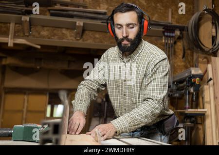 Horizontal photo serious carpenter with hearing protection operates a table saw, pushing a wooden board towards the rotating blade in a busy woodshop. Stock Photo