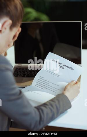 New job. Seen from behind modern 40 years old woman worker in modern green office in grey business suit with laptop and employee termination letter. Stock Photo