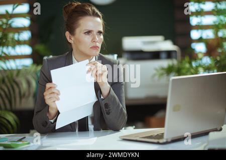 New job. pensive modern 40 years old woman worker in modern green office in grey business suit with laptop. Stock Photo