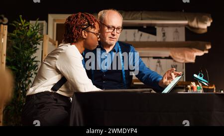 Craftsman teaching trainee to sew using textile industry tools and create fashion design. Beginner learning to manufacture clothing with experienced tailor in workshop. Handheld shot. Stock Photo