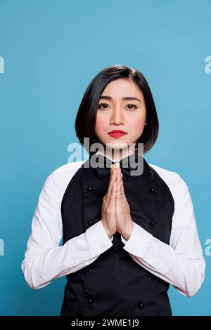 Young asian woman receptionist meditating with folded palms, relaxing and looking at camera. Attractive cafe waitress wearing uniform standing in zen pose portrait on blue background Stock Photo