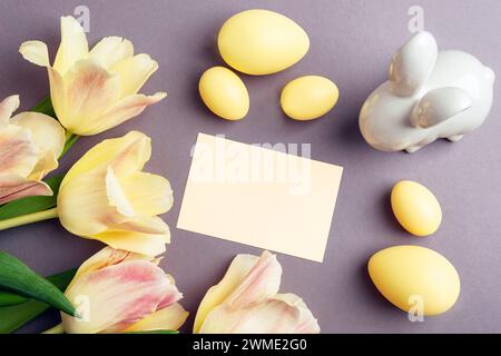 Easter background with blank card, yellow Easter eggs, bunny and tulip flowers on gray table. Top view, flat lay, mockup. Stock Photo