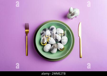 Table place setting with green plate, Easter eggs and bunny on purple background. Top view, flat lay, copy space. Stock Photo