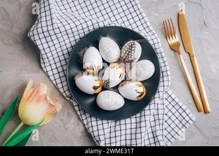 Easter eggs on black plate, checkered napkin and yellow tulip on concrete gray table. Top view, flat lay. Stock Photo