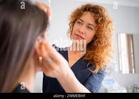 Female dermatologist performing procedure on woman customer. Cosmetologist with curly red hair make face care procedure. Focused aesthetic specialist do cosmetic procedure to client in beauty salon Stock Photo
