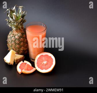 A mixture of freshly squeezed juices from tropical fruits on a black background, next to the halves of ripe grapefruit and pineapple. Close-up. Stock Photo