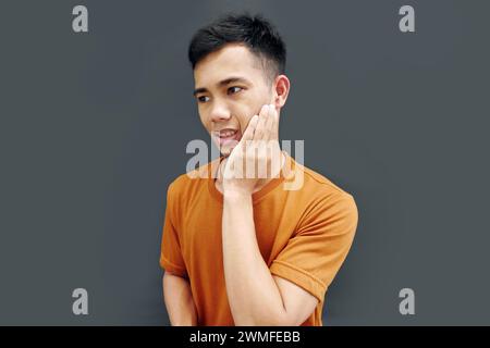 Grimacing young man having severe toothache, holding his cheek with hand, face in pain, isolated on copy space background Stock Photo