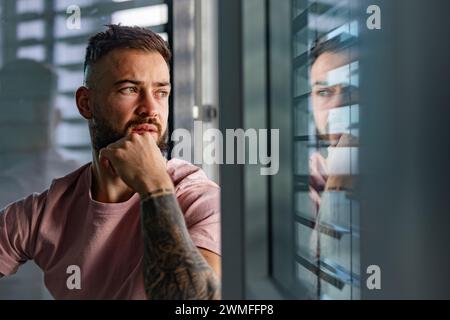 Handsome calm man in pink t-shirt with tattoo on arms looking through window. Stock Photo