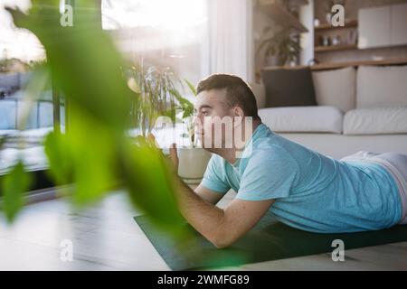 Young man with Down syndrome exercising at home, doing yoga, meditating. Man is praying, lying on belly. Stock Photo
