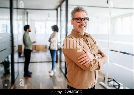 Senior businessman in casual wear stands with crossed arms in office, a blurred conversation between colleagues in the background, a moment of individual focus within a collaborative team environment Stock Photo