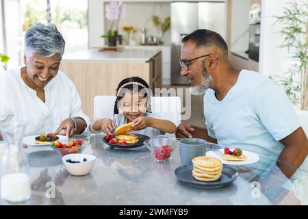 Biracial grandparents and granddaughter enjoy breakfast pancakes together at home Stock Photo