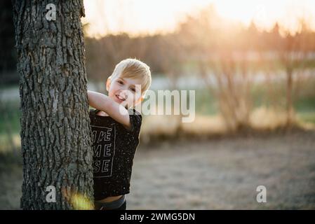 Close up of young boy smiling and peeking around tree in yard Stock Photo