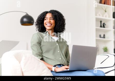 A cheerful African-American woman interacts with her laptop, her expression radiating confidence and ease in her home workspace. Female freelancer working remotely Stock Photo