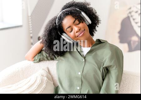 Eyes closed and with serene expression, the woman enjoys a moment of relaxation with her headphones on, embodying a peaceful break in a comfortable, modern setting. It's a snapshot of modern self-care Stock Photo