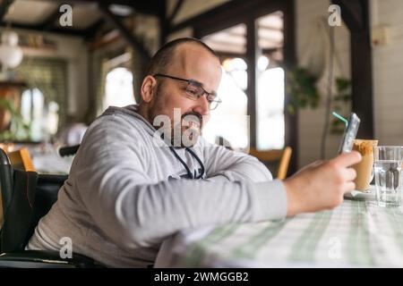 Happy paraplegic handicapped man in wheelchair is sitting at restaurant and using smartphone. Stock Photo