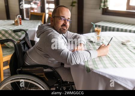 Happy paraplegic handicapped man in wheelchair is sitting at restaurant and using smartphone. Stock Photo