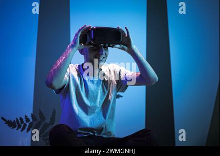 Young man wearing VR glasses sitting in a yoga pose on the virtual reality escape room floor. Fun, entertainment, and innovation concepts. Stock Photo
