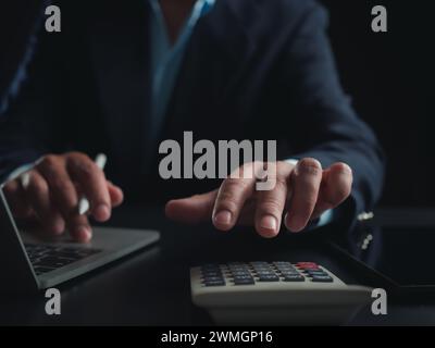 Businessman in black suit working on laptop computer and tablet and calculator, Hand Hands touch on tablet at office with dark background, Online work Stock Photo