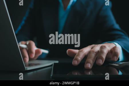 Businessman in black suit working on laptop computer and tablet, Hand Hands touch on tablet at office with dark background, Online working, Close up, Stock Photo
