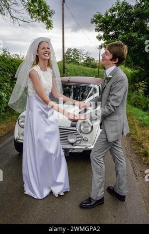 Old classic Mini Cooper car on street in English country side with bride and groom couple dancing before weeding ceremony. Stock Photo
