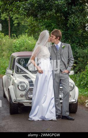 Old classic Mini Cooper car on street in English country side with bride and groom couple kissing  before weeding ceremony. Stock Photo