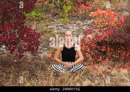 Happy young adult woman sitting in the lotus position against a background of autumn greenery and smiling. Ideal meditation. Stock Photo