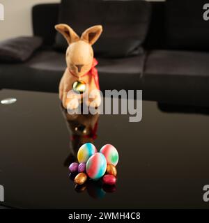 Group of easter eggs in different sizes and colors on table with blurry easter bunny in the background.  Square format. Stock Photo