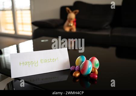 Group of easter eggs in different sizes and colors on table with blurry easter bunny in background and Happy Easter card in the foreground. Stock Photo