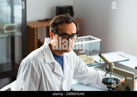 An adult male Caucasian scientist in glasses and a white coat is distracted from working with the sample being studied on a microscope and looking for Stock Photo