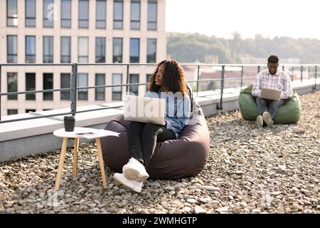 Successful business woman dressed in denim shirt and pants sitting at chair bag on roof top outdoors and typing on modern laptop, African American col Stock Photo
