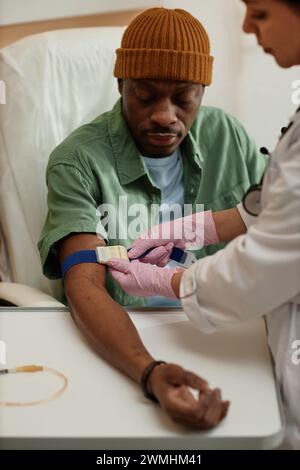 Vertical portrait of African American man preparing for IV treatment in clinic with nurse putting tourniquet on arm Stock Photo