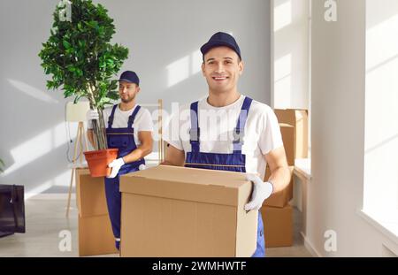 Young man mover worker in uniform holding a cardboard box and looking at camera in apartment. Stock Photo