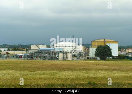 Large storage tanks and a network of pipes dominate the industrial landscape at an oil refinery under a cloudy sky. A field of tall grasses in the for Stock Photo