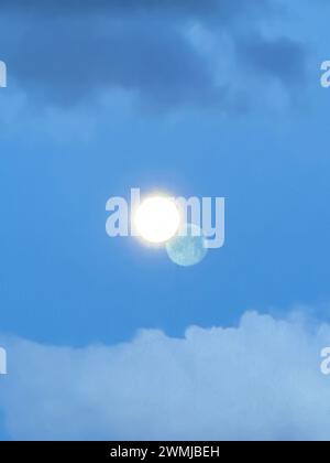 A bright moon shining against a blue sky with a shadow image in the distant clouds looks like a double moon with yellow circle or two distant planets. Stock Photo